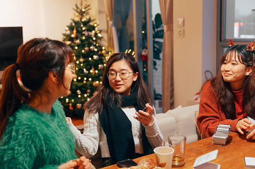 A half-length shot of three young smiling Asian women opening gifts or presents for Christmas while sitting around table talking with each other, enjoying time together.

Young Asian friends or family having fun together while gathering in a Christmas party at home indoors to celebrate Christmas.