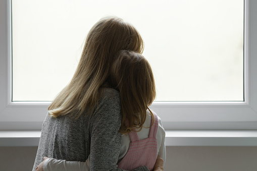 Young adult mother and daughter together looking out from window at home. Waiting, longing or sadness concept. Closeup. Back view.