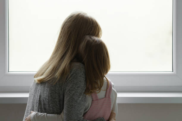Young adult mother and daughter together looking out from window at home. Waiting, longing or sadness concept. Closeup. Back view. stock photo