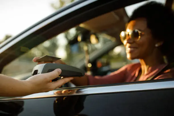 Close up shot of charming mid adult woman sitting in driver's seat of her car and using a credit card and paying for her order at the drive through or curbside pickup to the service person.