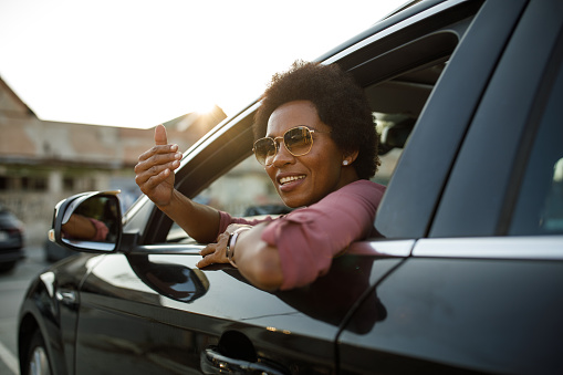 Portrait of charming mid adult African American woman peeking out of her car window, smiling at camera and gesturing you to join her for a ride.
