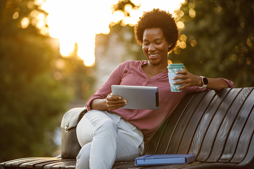 Portrait of charming mid adult African American businesswoman spending her coffee break relaxing on the street bench, enjoying takeaway coffee and using digital tablet.