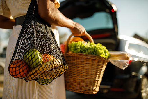 Rear view of unrecognizable woman carrying a mesh bag and a wooden basket with fresh produce from the farmer's market, walking towards the car, about to load it in the trunk.