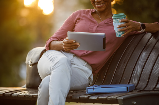 Cut out shot of charming mid adult African American businesswoman spending her coffee break relaxing on the street bench, enjoying takeaway coffee and using digital tablet.