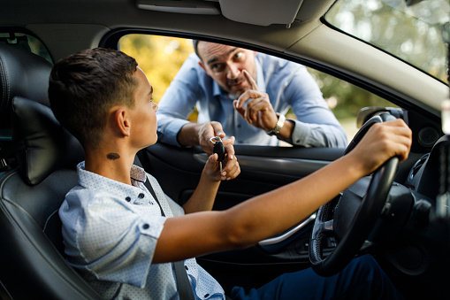 Candid shot of serious mid adult man giving car keys to his teenage son sitting behind the wheel, gesturing him to be careful when driving.