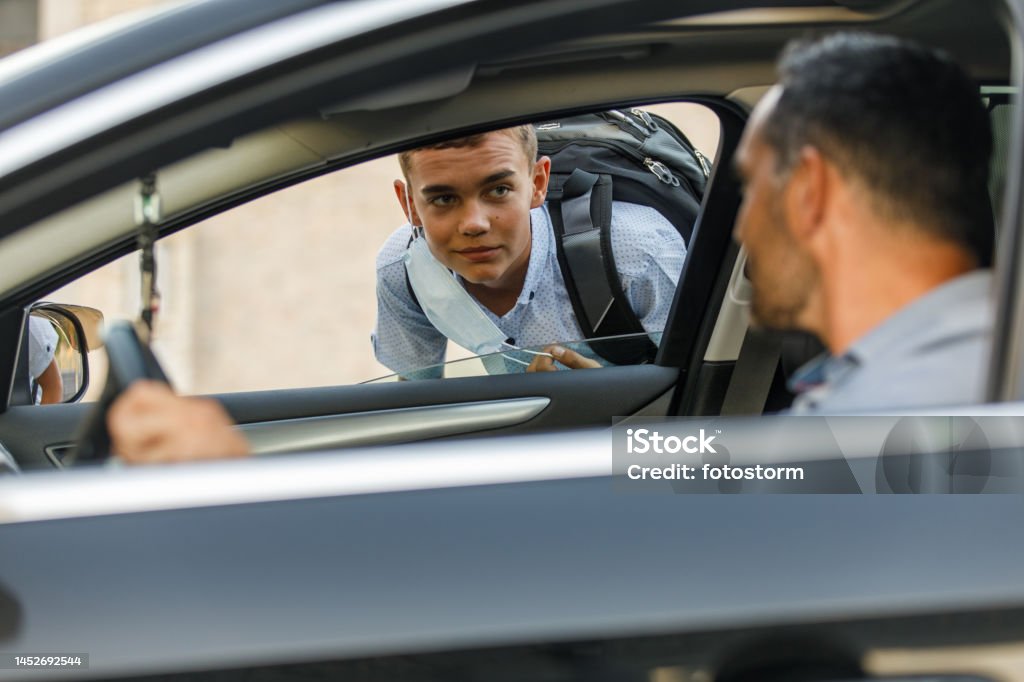 Man saying hi to his teeange son that he is picking up from school Selective focus shot of teenage boy greeting his father who is sitting behind the wheel, in his car, that came to pick him up from school. Child Stock Photo