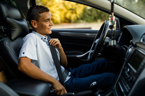 Copy space shot of smiling teenage boy sitting in driver's seat, putting on a seat belt before driving.