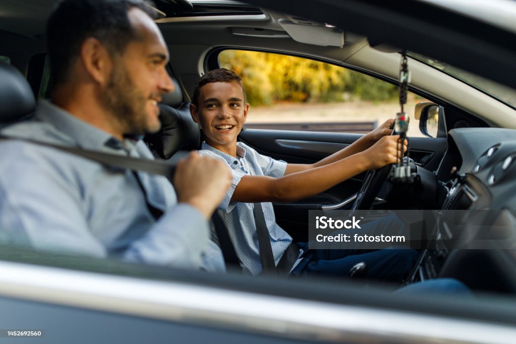 Man putting on seatbelt when teaching his teenage son to drive Selective focus shot of cheerful mid adult man sitting in passenger’s seat and putting on seatbelt when teaching his teenage son to drive. Driving Stock Photo