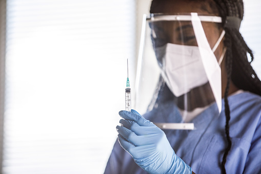 Copy space shot of young nurse, wearing disposable gloves, N95 face mask and face shield, preparing a vaccine or a shot in a syringe.