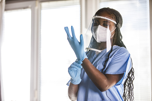 Copy space shot of unrecognizable Black nurse putting on disposable protective gloves before examining a patient. Part of a series. She is wearing N95 face mask and face shield.