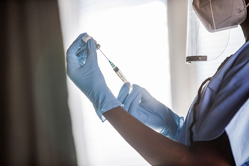 Cut out shot of young nurse, wearing disposable gloves, N95 face mask and face shield, preparing a vaccine or a shot in a syringe. She is drawing medicine from a vial.