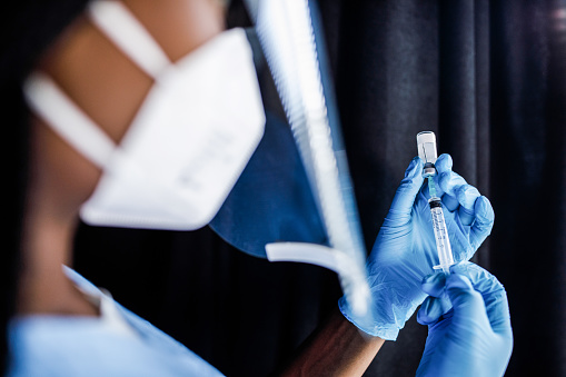 Over the shoulder view of unrecognizable nurse, wearing disposable gloves, drawing a vaccine or a shot in a syringe from a vial.