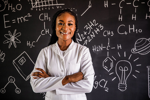 Portrait of confident female scientist, in a lab coat, standing, with arms crossed, in front of a blackboard with written chemistry formulas. She is looking at camera and happily smiling.