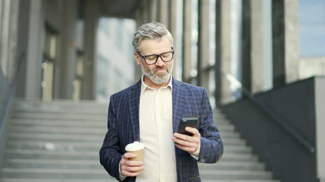 Confident mature businessman with cup of coffee hold smartphone scrolling browsing texting at modern business centre