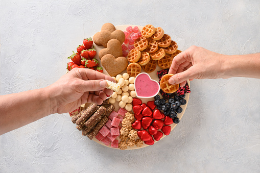 Male and female hands take chocolate sweets and treats from Valentines Day charcuterie board on light gray background. View from above.
