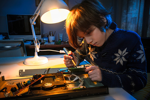 Little genius boy using a soldering iron to fix an electronic device at the desk in the room.