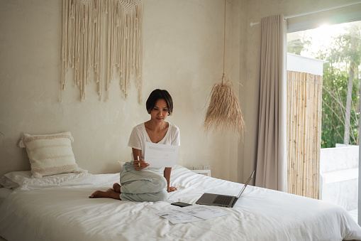 A mid-adult Thai woman is sitting on a bed in bedroom and looking at tax documents.