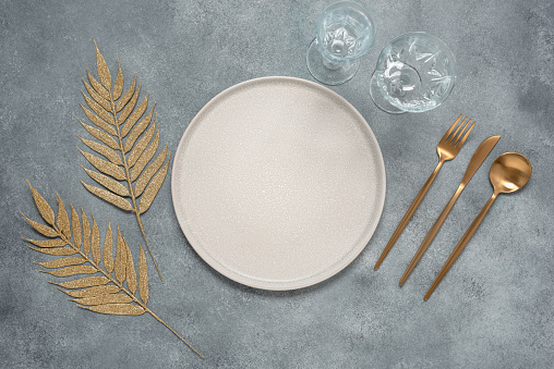 Christmas or New Year minimal table setting. Empty beige plate, golden cutlery and golden leaves on a gray concrete background. Top view, flat lay, selective focus.