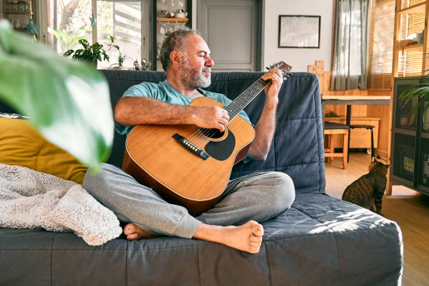 istock Middle-aged bearded man playing acoustic guitar while sitting on sofa in light living room. 1452719462