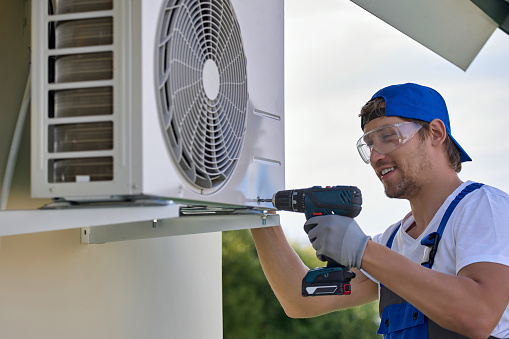 Smiling caucasian worker installs the outer metal case of the air conditioner with a screwdriver. Qualified male installer in uniform fixes air conditioner mount with tool