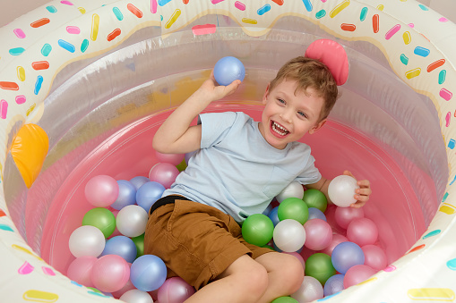 Smiling boy is having fun playing at home with colorful balloons. Happy Caucasian child throws plastic balls, lying in dry pool. Top view of the children's dry pool for fun games.