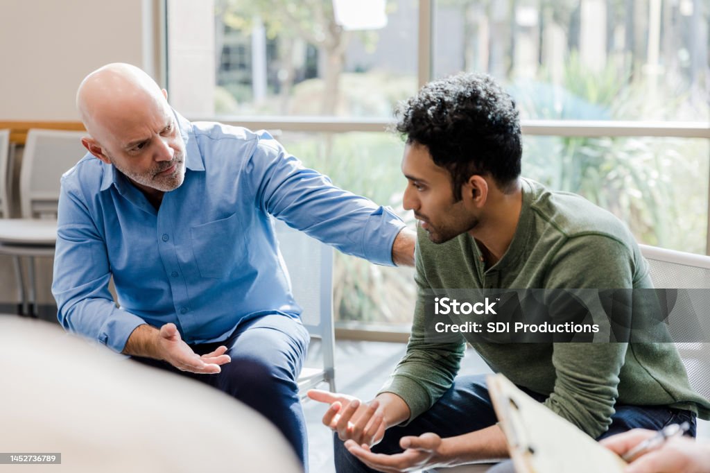 Mature man helps younger man verbalize problems in therapy The empathetic mature adult man gestures and asks questions as he helps the younger man work through his problems. Mental Health Stock Photo