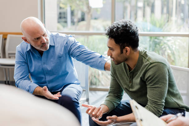 Mature man helps younger man verbalize problems in therapy The empathetic mature adult man gestures and asks questions as he helps the younger man work through his problems. mental health stock pictures, royalty-free photos & images