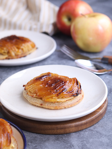 A traditional mini French thin apple tartlet on a black plate with it fruits, fork and spoon in background. A dessert made with puff pastry pie, caramelized with brown sugar.