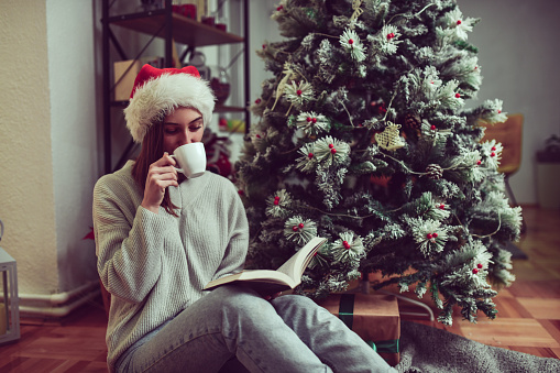 Cute Female With Santa Hat Enjoying Coffee Time While Reading Book Under Christmas Tree