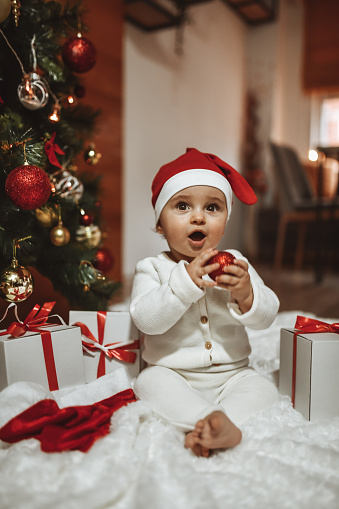 Baby Playing Santa Claus Under Christmas Tree And Holding Bauble In Hand