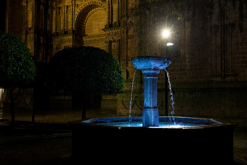 Plasencia, Spain - December 25, 2022: Rainy night in the city of Plasencia, with the Fuente del Cabildo in the foreground and part of the Cathedral of this city in the background.