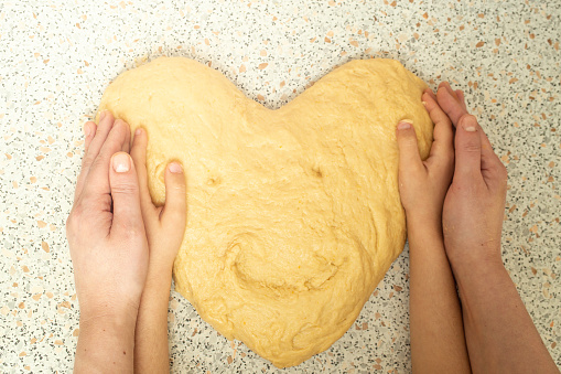 Mother and son hands holding dough in the shape of a heart with a smiley face, soft focus close up