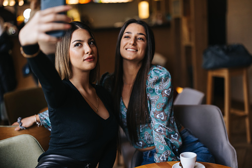 Two young cheerful female friends taking selfie in cafe