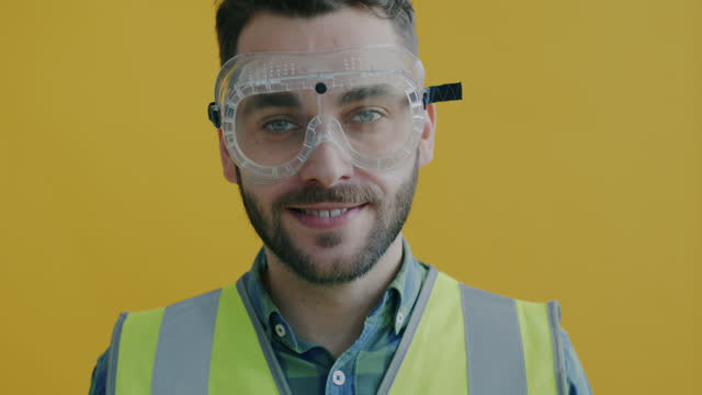 Slow motion portrait of young man in safety vest putting on industrial goggles and smiling on yellow background
