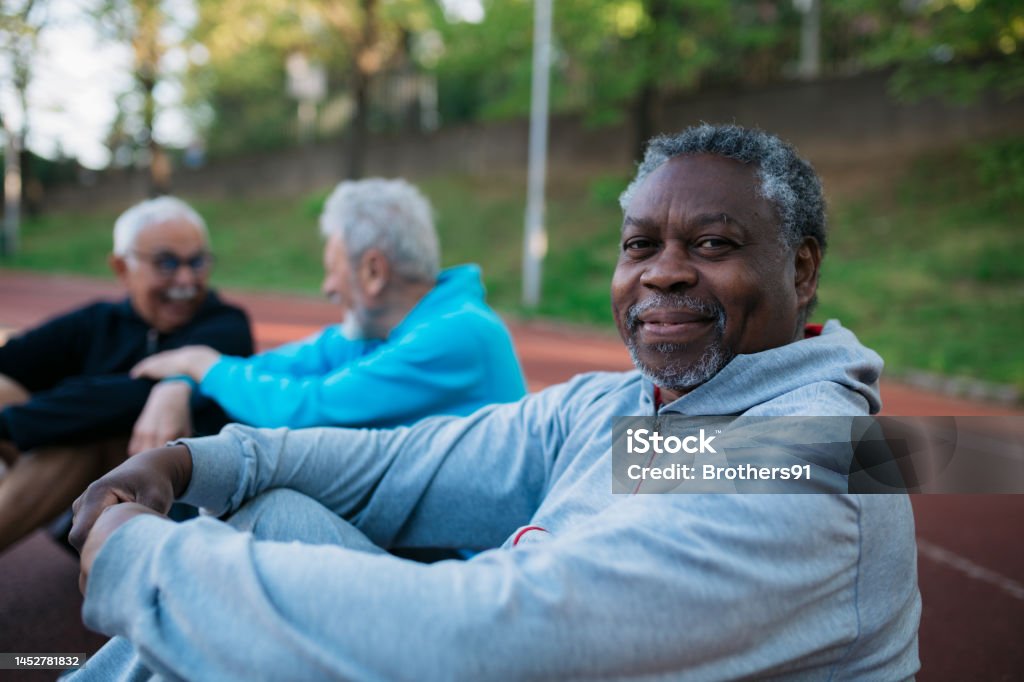 Senior man taking rest after workout at outdoors exercise court Portrait of a healthy elderly man relaxing on a running track with friends sitting next to him. Senior man taking rest after workout at outdoors exercise court. Men Stock Photo