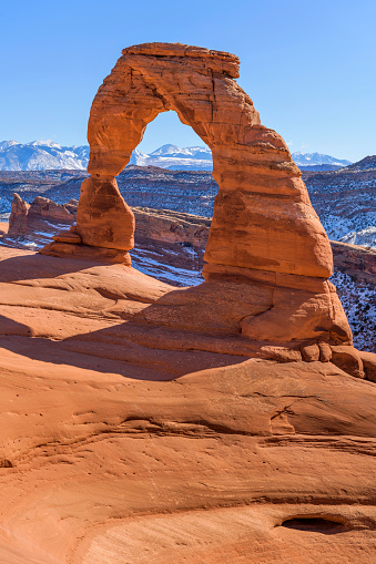 A closeup vertical view of Delicate Arch, with snow-covered La Sal Mountains towering in background, on a clear sunny Winter day. Arches National Park, Utah, USA.