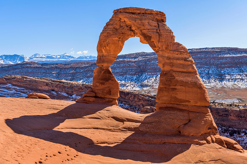 A closeup view of Delicate Arch, with snow-covered La Sal Mountains towering in background, on a clear sunny Winter day. Arches National Park, Utah, USA.