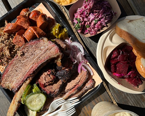 An overhead view of a large tray of Texas barbecue foods, including beef rib, beef brisket, sausage, pulled lamb, plus sides of pickles, mac and cheese, grits, beets, bread, and more.