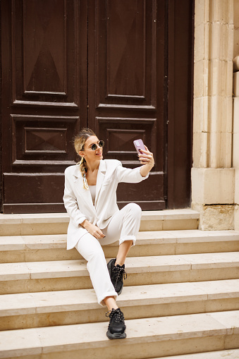 Beautiful young female fashion influencer sitting on staircase and taking selfie for social media outdoors