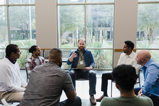Sitting in a circle, the diverse group of men listen attentively to the mid adult male therapist.