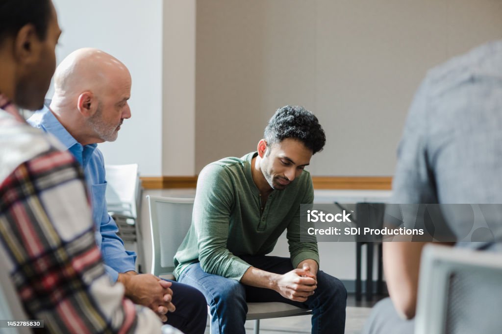 In group therapy, young man looks down while gathering thoughts During group therapy, the young adult man looks down at the floor as he takes time to gather his thoughts. Recovery Stock Photo