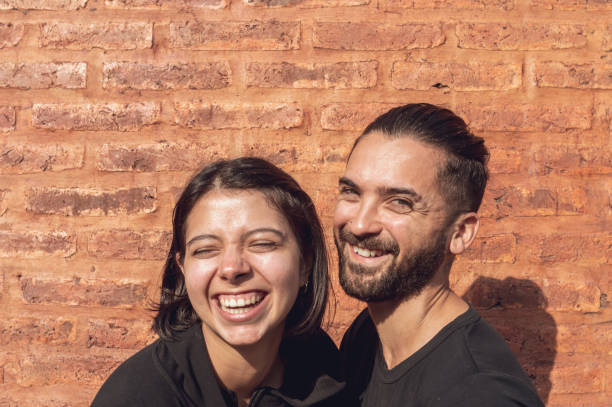 A cheerful couple laughing while looking at the camera against a brick wall background stock photo