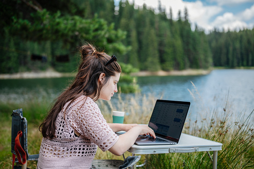 Young woman working with laptop during her camping vacation in mountain.Balancing between leisure and work.