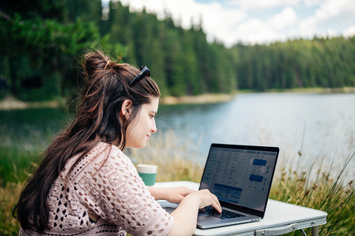 Shot of smiling young woman using laptop on camping vacation in the mountains for working remotely.