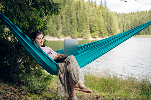 Young woman lying in hammock using a laptop for working remotely on a vacation.