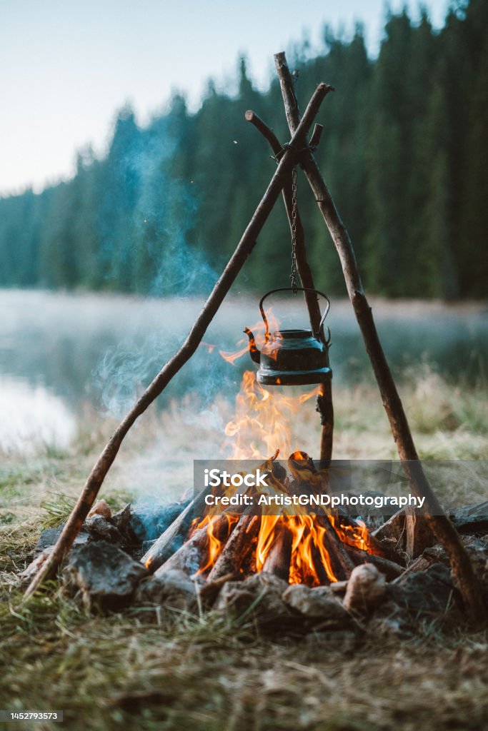 Peaceful atmosphere around the cozy campfire. Shot of the peaceful cozy campfire with a vintage teapot and beautiful lake. Camping Stock Photo