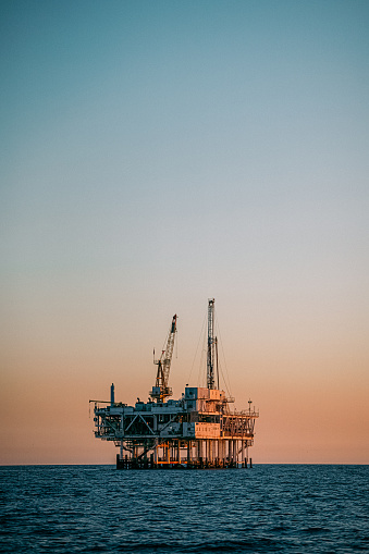 A beautiful photograph of an offshore oil drilling rig at sunset in Huntington Beach, California. The orange hues of the setting sun highlight the industrial machinery and equipment used in the drilling and extraction of fossil fuels, including crude oil and natural gas. \n\nThis image captures the intersection of the energy industry and the natural beauty of the Pacific Ocean, and speaks to issues of fuel and power generation, energy crises, and environmental concerns surrounding the oil and gas industry.
