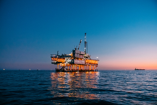 A beautiful photograph of offshore oil drilling at sunset in Huntington Beach, California. The orange and pink hues of the setting sun highlight the industrial machinery and equipment used in the drilling and extraction of fossil fuels, including crude oil and natural gas. A cargo ship is visible in the background.

This image captures the intersection of the energy industry and the natural beauty of the Pacific Ocean, and speaks to issues of fuel and power generation, energy crises, and environmental concerns surrounding the oil and gas industry.
