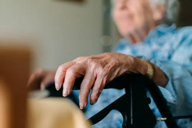 Close-Up Focus on Foreground Shot of an Unrecognizable Senior Woman Resting Her Hand on the Grip of her Mobility Walker while Sitting and Looking Out the Window