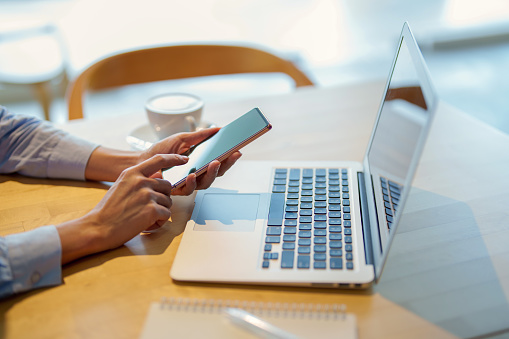 Woman's hands on a smart phone in front of a laptop close up, working at cafe. Technology and flexible working.